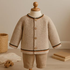 Könnte beinhalten: Beige Strickjacke und Hose für ein Kind. Die Strickjacke hat einen runden Ausschnitt, lange Ärmel und Holzknöpfe. Die Manschetten und der Halsausschnitt sind mit schwarzen und orangefarbenen Streifen versehen. Die Hose ist einfarbig beige.