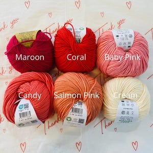 May include: Six balls of yarn in various shades of pink and red, including maroon, coral, baby pink, candy, salmon pink, and cream. Each ball has a label with the yarn color name and information about the yarn.