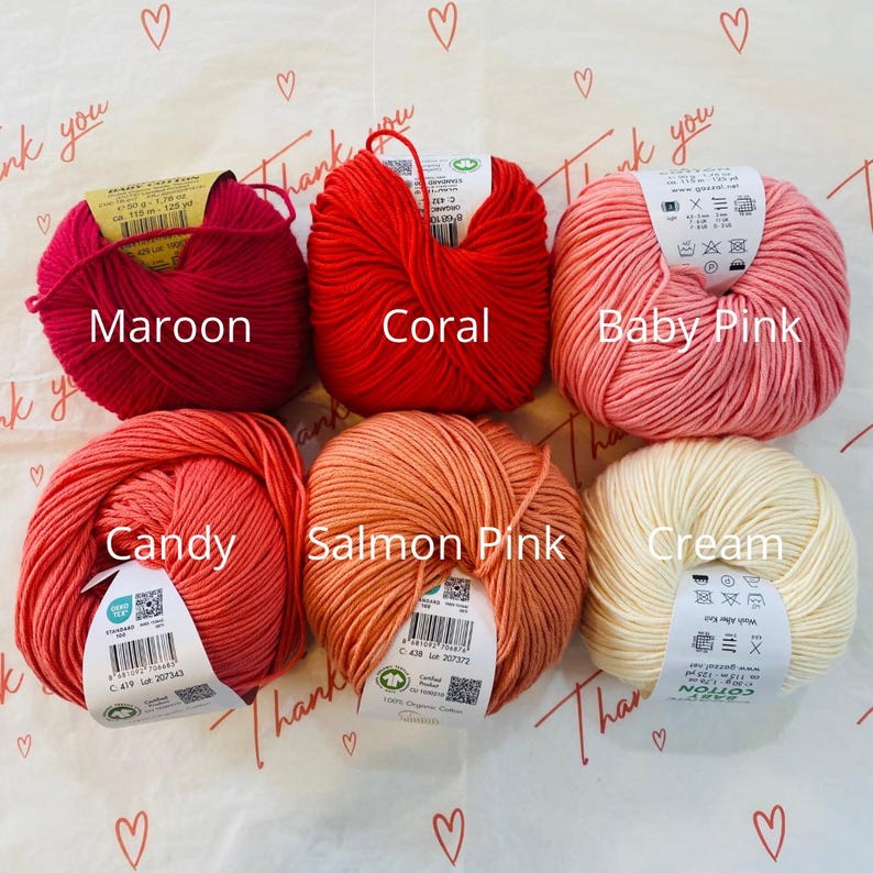 May include: Six balls of yarn in various shades of pink and red, including maroon, coral, baby pink, candy, salmon pink, and cream. Each ball has a label with the yarn color name and information about the yarn.