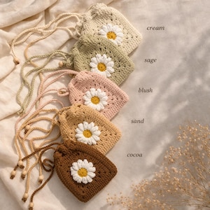 May include: Five crochet drawstring pouches arranged in a vertical row, each featuring a central white and yellow daisy motif. The pouches are colored cream, sage, blush, sand, and cocoa, with matching drawstrings and small wooden beads at the ends.