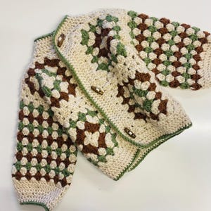 May include: A crocheted cardigan featuring a brown, green and white granny square pattern. The cardigan has a button fastening and long sleeves.