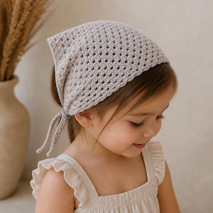 Puede incluir: Un pañuelo para la cabeza de estilo bandana de ganchillo gris claro, con forma cuadrada y cierre de lazo. El cubrecabezas está hecho de un material texturizado de tejido abierto. El pañuelo lo lleva una niña pequeña, combinado con un vestido de color claro.