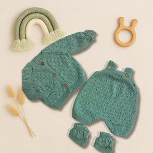 May include: A sage green knitted baby set featuring a long-sleeve button-up cardigan, a pair of overalls with button straps, and two matching booties. The set is accompanied by a decorative macrame rainbow and a light brown wooden ring toy.
