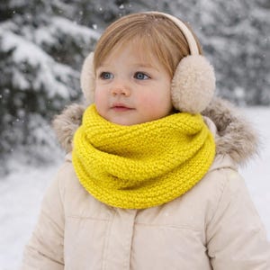 Peut inclure: Un enfant portant des accessoires d'hiver dans un paysage enneigé. L'enfant porte une écharpe tubulaire jaune vif, des cache-oreilles crème et une veste beige avec une capuche bordée de fourrure. L'arrière-plan montre une scène hivernale floue.