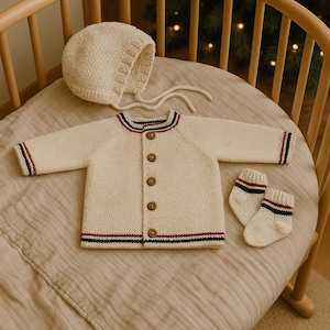 May include: A cream knitted baby cardigan with blue and red stripes, a matching cream knitted hat, and a pair of cream knitted booties with blue and red stripes.