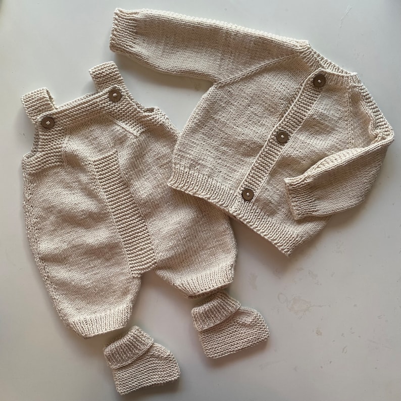 May include: A knitted baby outfit set in a cream color. The set includes a cardigan with button closures, overalls with a bib, and booties.