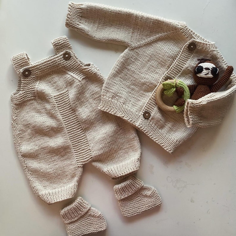 May include: A knitted baby outfit in a cream color, featuring a cardigan with button closures and a pair of overalls with foot coverings. The outfit is laid out on a white surface.