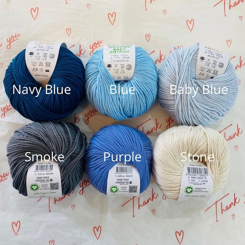May include: Six balls of organic cotton yarn in various colors: navy blue, blue, baby blue, smoke, purple, and stone. Each ball has a label with the brand name, yarn weight, and care instructions.