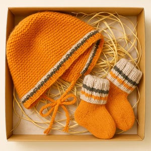 Op de afbeelding: Een oranje gebreide baby set, bestaande uit een muts en sokken. De muts heeft oorflappen en een strik. Zowel de muts als de sokken hebben grijze en gebroken witte strepen. De set wordt gepresenteerd in een kartonnen doos.