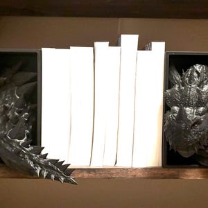 May include: Black dragon head bookends holding white books on a wooden shelf. The bookends are detailed with textured scales and horns, adding a fantasy theme to the bookshelf. The dragon heads are positioned on either side of the books.