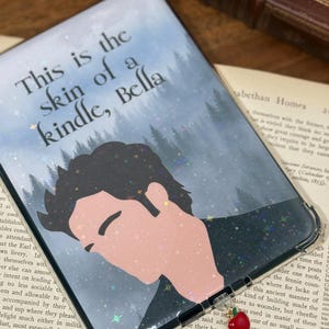 May include: Clear Kindle case with a design featuring a silhouette of a person and the text "This is the skin of a kindle, Bella." The case has a glittery, starry background and a red apple charm.