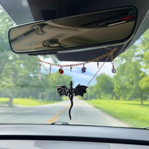 May include: A car rearview mirror charm featuring a black dragon pendant suspended from a gold-toned chain. The chain is adorned with several small charms, including a sword, a red sphere, a key, a purple flower, a lightning bolt, and crystals.