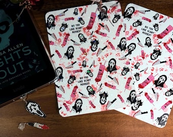 May include: Two white decorative coasters with a repeating pattern of a ghost face, knives, and pink cell phones. The text "DO YOU LIKE SCARY MOVIES?" is also printed on the coasters. A Kindle e-reader and a beaded charm are also visible.