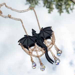 May include: A black dragon pendant necklace with gold chain and crystal accents. The dragon is in flight with its wings spread wide. The necklace is hanging from a mirror.