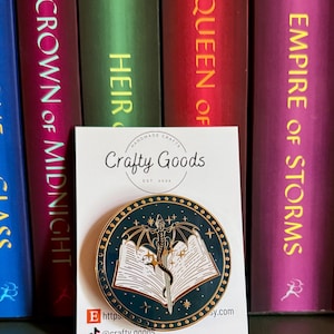 May include: A round enamel pin with a dragon and open book design, set against a dark teal background with gold accents. The pin features the text "Crafty Goods" and social media handles. The pin is displayed in front of a row of colorful books.