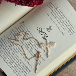 May include: A gold-colored necklace with a sword pendant and a white dragon pendant rests on an open book. The book's pages display text, with a red rose in the background. The chapter title "THIRTY-ONE" is visible.