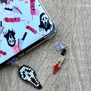 May include: A clear phone case with a horror-themed pattern featuring a white ghost face, knives, and blood splatters. Two beaded charms hang from the phone case: a black and white ghost face and a red and silver knife.