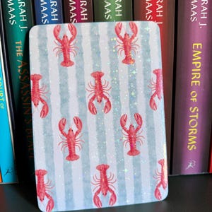 May include: Rectangular decorative item with a light blue and white striped background, featuring red lobster illustrations. The item has a glossy finish and is positioned in front of a row of books.