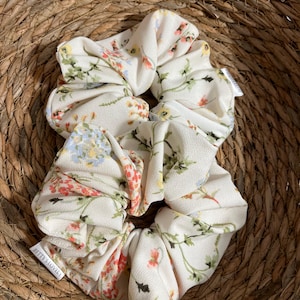 May include: Two ivory scrunchies with a delicate floral pattern in shades of red, blue, and green. The scrunchies are made of a soft fabric and are labeled "Betty's Lovely". They are resting in a woven basket.