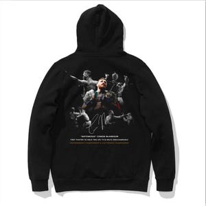 May include: Black hooded sweatshirt with a graphic print of Conor McGregor, a mixed martial arts fighter, with the text "NOTORIOUS CONOR MCGREGOR" and "FIRST FIGHTER TO HOLD TWO UFC TITLE BELTS SIMULTANEOUSLY FEATHERWEIGHT CHAMPIONSHIP & LIGHTWEIGHT CHAMPIONSHIP"