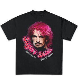 May include: Black t-shirt featuring a graphic of Sean O'Malley with pink hair. The design includes the text "Sugar Sean" in a pink and white font, along with "12 Knockouts" and "Sean O'Malley".