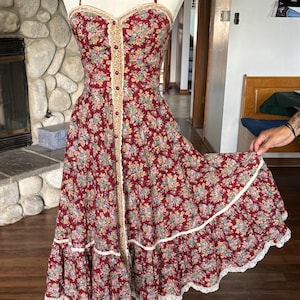 May include: A sleeveless midi dress in a deep red fabric featuring a multi-colored floral pattern. The dress has a sweetheart neckline, thin shoulder straps, a central button-down front, and a tiered skirt hem accented with cream-colored lace trim.