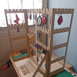 Puede incluir: Un expositor de madera con varios estantes para exhibir llaveros. El expositor está hecho de madera marrón claro y tiene un acabado natural. Los estantes están dispuestos en forma de cuadrícula y tienen pequeños agujeros para colgar llaveros. Hay varios llaveros colgados de los estantes, incluyendo una mariposa, una huella de pata, una estrella y un círculo. El expositor está sobre una alfombra de corte verde.
