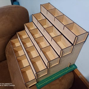 May include: A stepped wooden organiser with numerous rectangular compartments. The organiser is made of light-coloured wood with dark brown edges. It is designed to hold and display small items, suitable for craft supplies or collectibles.