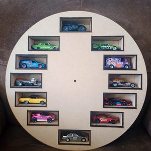 Pode incluir: Uma prateleira circular de madeira com doze compartimentos retangulares, cada um contendo um carro de brinquedo em miniatura. Os carros são de várias cores, incluindo azul, verde, amarelo, rosa e vermelho. A prateleira é de madeira castanha clara.