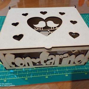 May include: A wooden box with a heart-shaped cutout and two birds inside. The box is decorated with the words "Con Cariño" and several hearts.