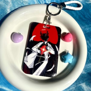 Könnte beinhalten: Eine Schwarzweißillustration einer Person mit Hörnern und einem Heiligenschein, die sich an die Brust fasst. Die Illustration ist auf rotem Hintergrund und auf einem weißen Plastik-Schlüsselanhänger gedruckt.