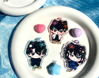May include: Three black and white cartoon character keychains with pink, blue, and white accents. The characters have black hair, black eyes, and are wearing different outfits. The keychains are on a white plate with a blue background.