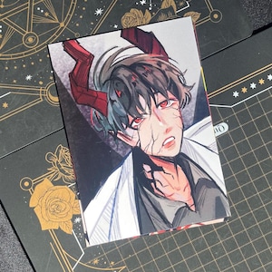 Könnte beinhalten: Ein rechteckiger Kunstdruck mit einer detaillierten Illustration eines Charakters mit roten Augen und Hörnern. Der Charakter hat dunkles Haar und sichtbare Gesichtsverletzungen. Der Druck ist vor einem dunklen Hintergrund mit anderen dekorativen Karten platziert.