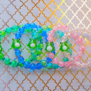 May include: Five colorful beaded bracelets with frog charms. The bracelets are made with blue, green, and pink beads. The frog charms are green with white eyes.