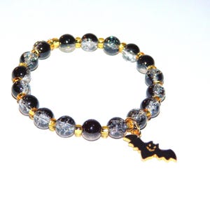 May include: A beaded bracelet featuring alternating black beads and clear crackle-textured beads, separated by small gold-toned spacers. A small, black, gold-edged bat-shaped charm hangs from the bracelet, which is arranged in a circle.