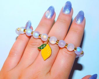 May include: A beaded bracelet featuring translucent, iridescent spherical beads alternating with small gold-toned metallic spacers. A single yellow lemon-shaped charm with a green leaf detail hangs from the center of the elasticized beaded strand.