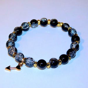 May include: A beaded bracelet featuring black and silver-speckled beads, interspersed with gold-colored spacers. A gold-colored arrow charm with a heart detail hangs from the bracelet. The bracelet is displayed against a light blue background.