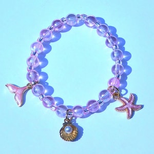 May include: A pink beaded bracelet with three charms: a pink mermaid tail, a gold seashell with a pearl, and a pink starfish. The bracelet is made of clear, iridescent beads and is on a light blue background.