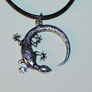 May include: A silver-toned lizard pendant necklace with a black leather cord. The lizard is curled into a circle with its tail forming the top of the circle. The lizard is decorated with a geometric pattern.