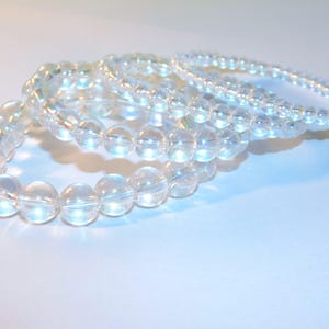 May include: Four clear quartz crystal bead bracelets. The bracelets are made of round, translucent beads strung together. The beads reflect light, creating a shimmering effect. The bracelets are arranged in a slightly overlapping pattern, suggesting they can be worn together.
