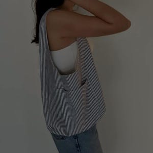 May include: A blue and white striped tote bag with a large front pocket. The bag is made of a lightweight fabric and has a casual, relaxed style. The bag is worn over the shoulder.