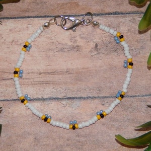 May include: White beaded bracelet with small yellow and blue flower accents. The bracelet has a silver clasp.