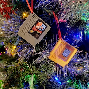 May include: Two vintage-style video game cartridge ornaments hanging on a Christmas tree. The ornaments are decorated with the text "Super Mario Bros." and "Tetris".