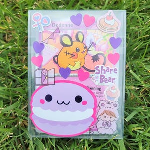 May include: A clear plastic case filled with stickers and a card. The card features an orange and yellow cartoon mouse character. Stickers include purple and pink hearts, a macaron, and a girl in a bear costume.
