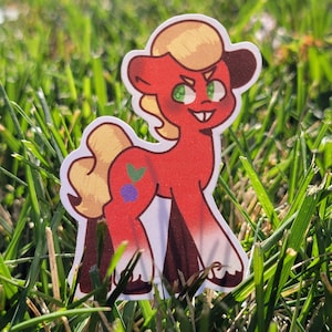 Könnte beinhalten: Ein rotes Comic-Pferd mit einer blonden Mähne und einer blauen Beere an der Seite. Das Pferd steht auf einer grünen Grasfläche.