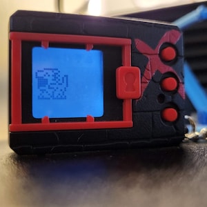 May include: A black and red handheld digital pet device with a blue screen showing a pixelated character. The device has red buttons and accents, and a silver chain. The device is on a dark surface.