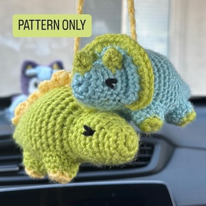 May include: Two crocheted dinosaur ornaments in shades of green and blue, with yellow accents. The dinosaurs have black eyes and are hanging from a yellow string. The text "PATTERN ONLY" is in a green box.