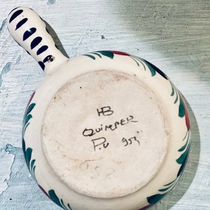 May include: White ceramic bowl with a green and red floral design. The bowl has a handle and is marked with the text "HB Quimper P. 357".