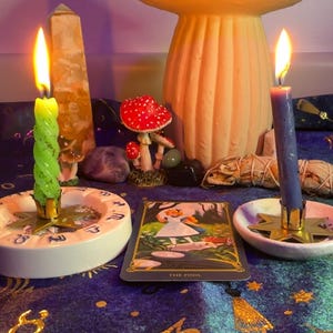 Könnte beinhalten: Eine mystische Szene mit brennenden Kerzen, einer pilzförmigen Lampe und einer Tarotkarte. Die Kerzen sind grün und blau, mit Flammen. Die Tarotkarte zeigt "Der Narr". Die Szene spielt sich auf einem dunkelblauen Tuch mit goldenen Sternmustern ab.