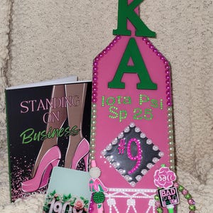May include: Pink and green decorative items, including a paddle with "AKA" and "Spotlight" text, a book titled "Standing on Business", a pen, and a bracelet. The paddle has pearl and rhinestone accents.